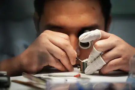 A jeweler meticulously works on a small, intricate silver watch component using precision tools.