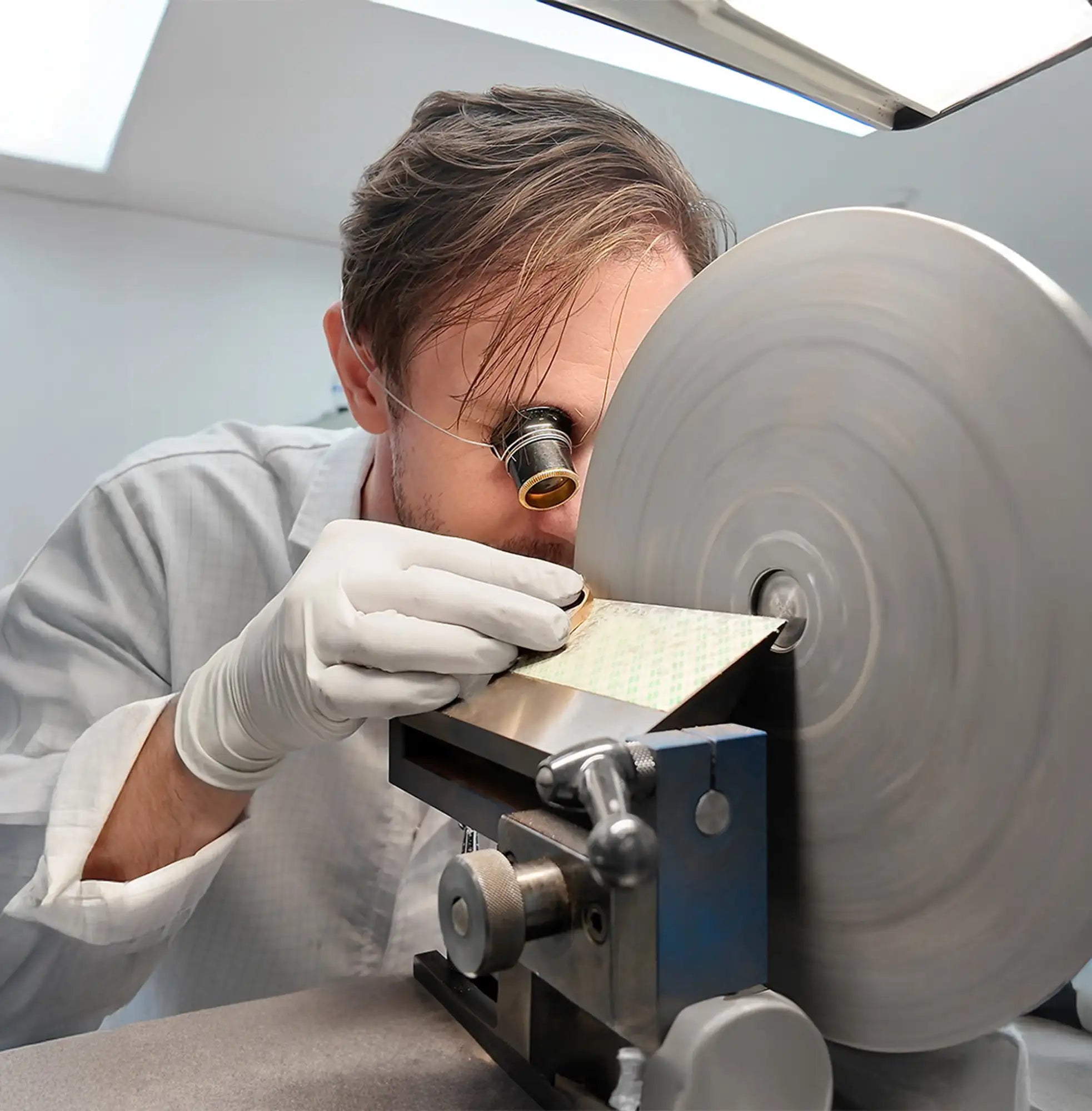 A man in a white lab coat and gloves carefully examines a small, rectangular piece of gold-colored metal being polished by a rotating grinding wheel.
