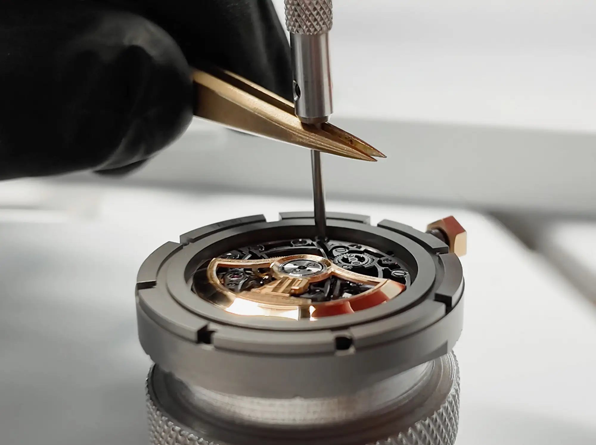A precision-engineered watch movement with intricate gold-toned gears and a dark gray case, being meticulously adjusted by a jeweler’s tool.