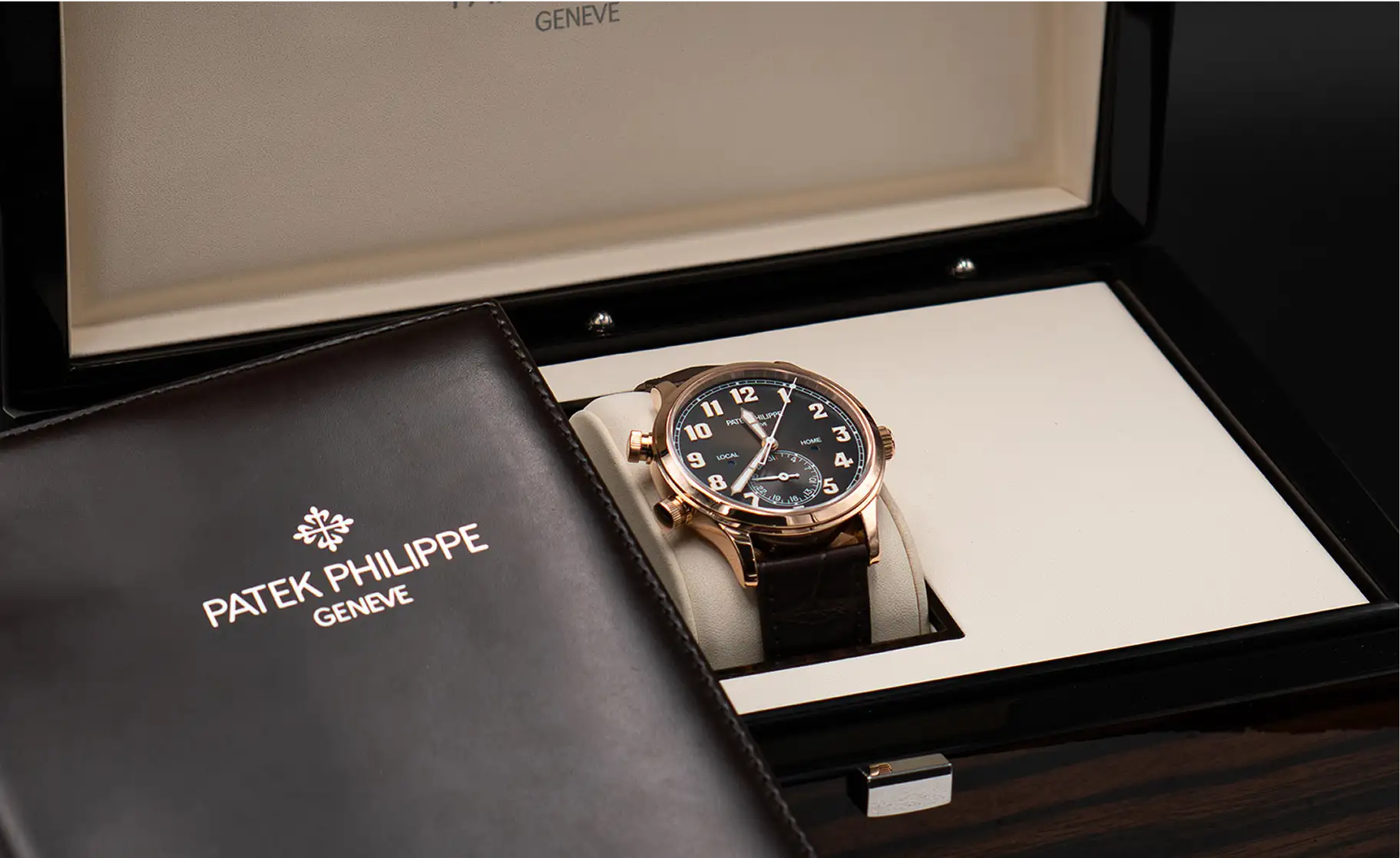 A rose gold patek philippe geneve watch with a black dial and dark brown leather strap, nestled in its presentation box.
