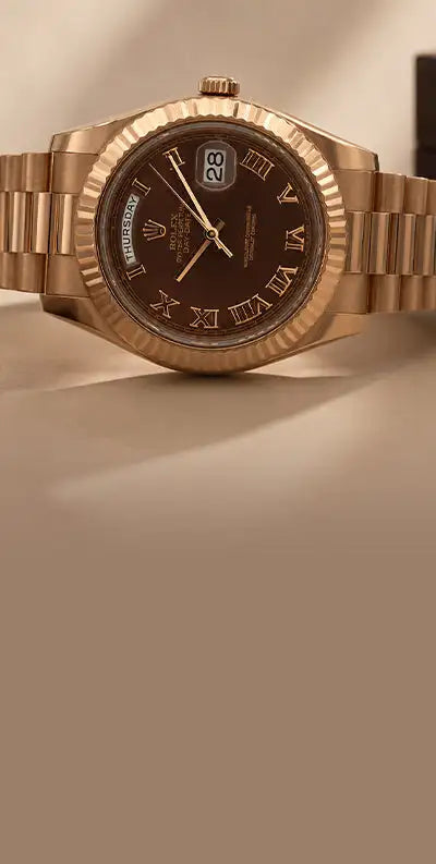 A rose gold rolex day-date watch with a dark brown dial, roman numeral hour markers, and an integrated bracelet.