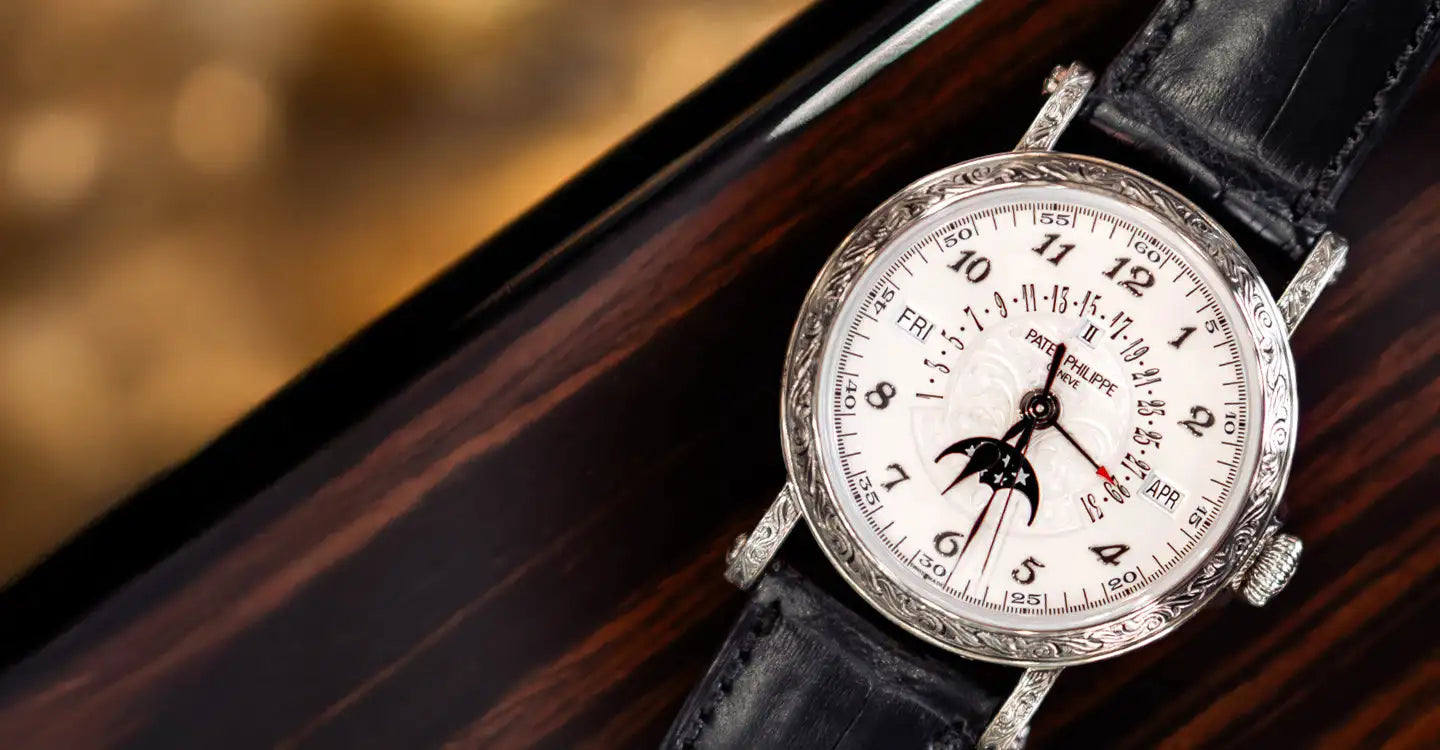 A silver-toned patek philippe watch with a white dial, black leather strap, and intricate engraved bezel.