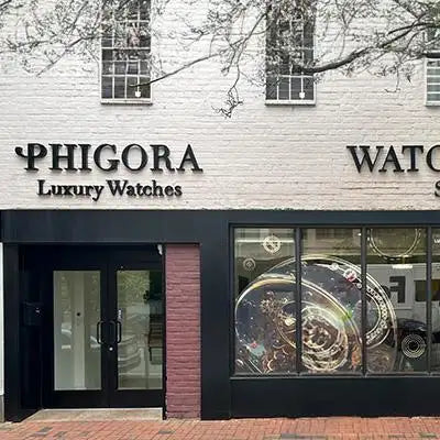 The storefront of phigora luxury watches, featuring a white brick facade with black lettering and large display windows showcasing intricate watch mechanisms.